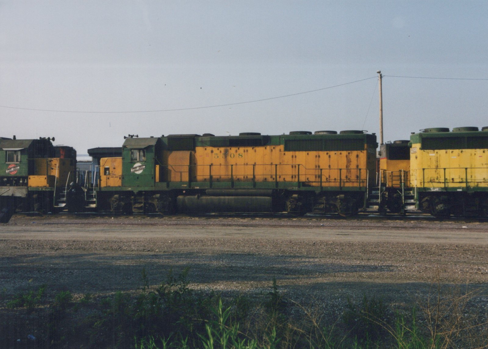 A side view of a green and yellow Chicago and North Western diesel locomotive, number 5508, parked in a gravel rail yard with other locomotives behind it under an overcast sky.