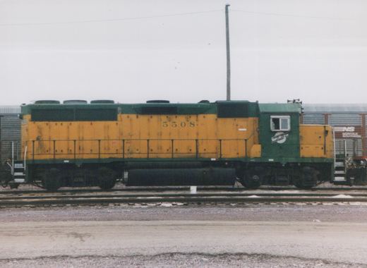 A side view of a green and yellow Chicago and North Western diesel locomotive, number 5508, in a yard with a Norfolk Southern boxcar in the background.