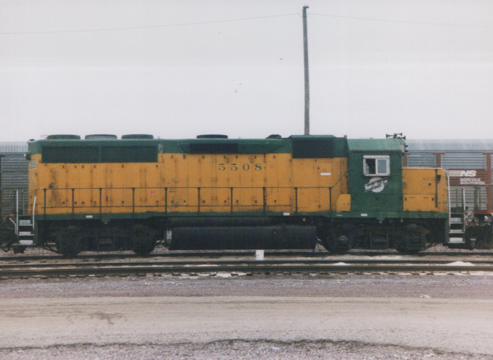 A side view of a green and yellow Chicago and North Western diesel locomotive, number 5508, in a yard with a Norfolk Southern boxcar in the background.