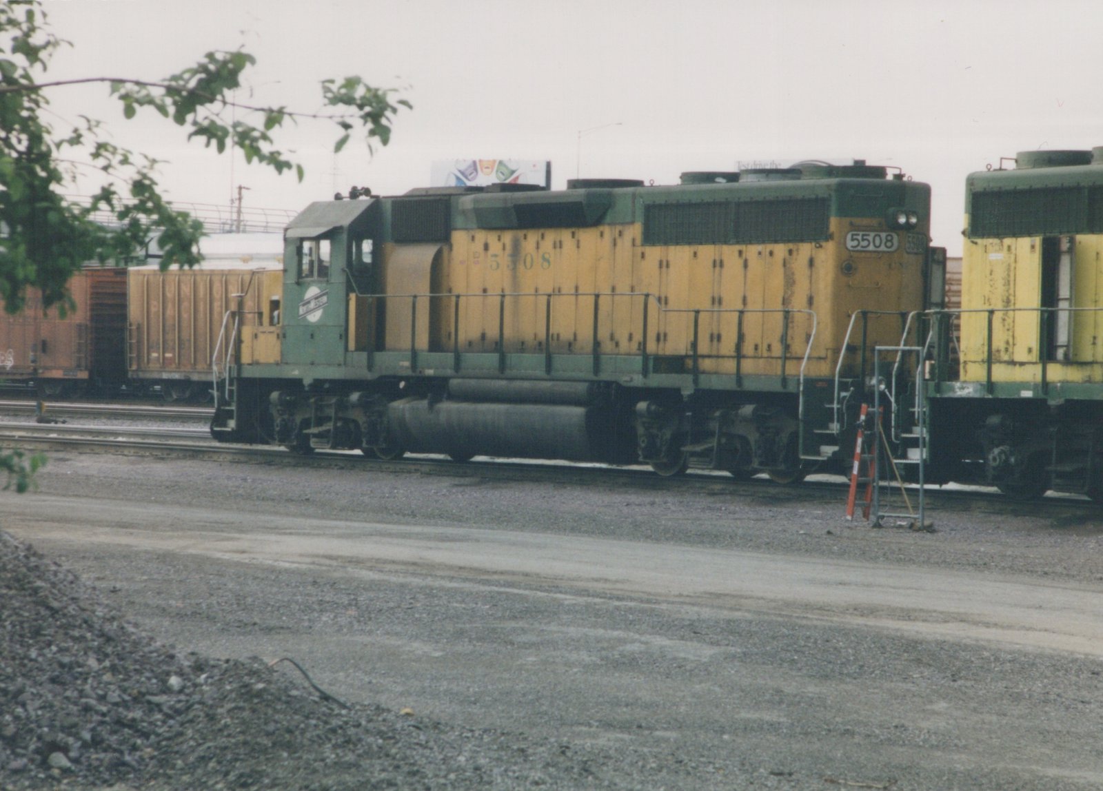 A green and yellow Chicago and North Western diesel locomotive, number 5508, is parked on tracks in a gravel yard with a boxcar in the background.