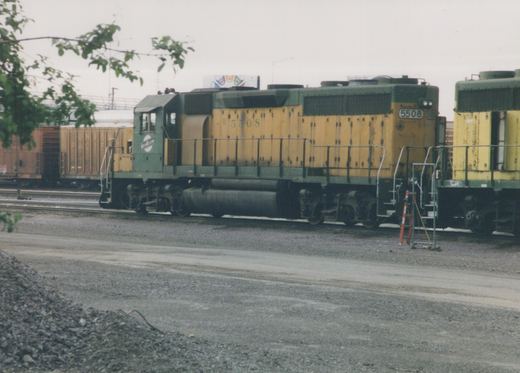 A green and yellow Chicago and North Western diesel locomotive, number 5508, is parked on tracks in a gravel yard with a boxcar in the background.