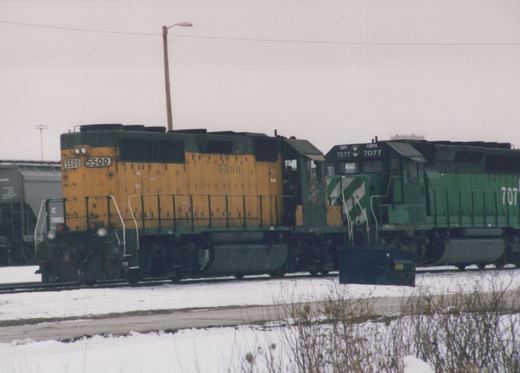 A yellow Illinois Central locomotive #5500 and a green CEFX locomotive #7077 stand next to each other on snow-covered tracks in a railway yard.