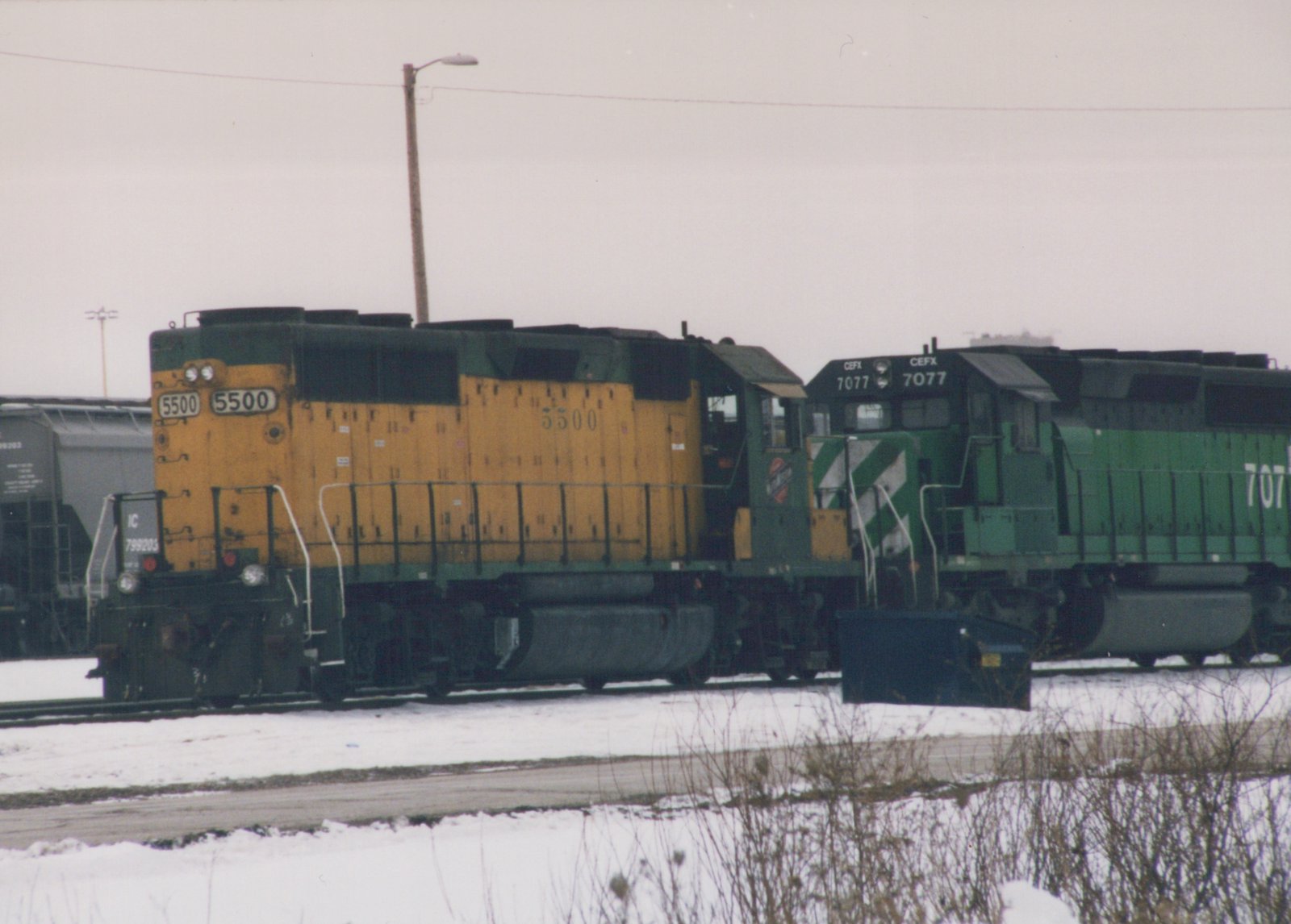 A yellow Illinois Central locomotive #5500 and a green CEFX locomotive #7077 stand next to each other on snow-covered tracks in a railway yard.