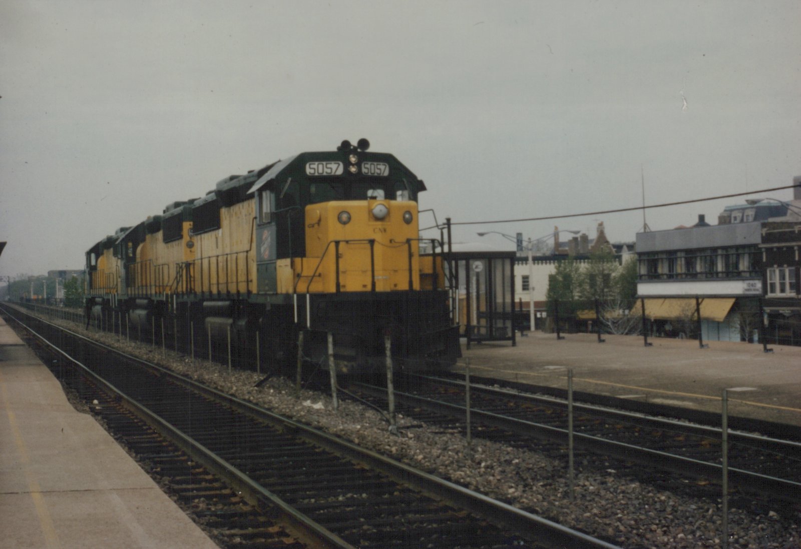 A yellow and black Chicago and North Western diesel locomotive with the number 5057 is stopped at a station platform next to a modern shelter.