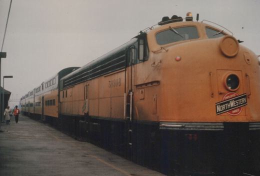 A side-view of a light brown and black diesel locomotive with 'NORTHWESTERN' on the front and number 5030B on the side, stopped at a station platform with two people walking nearby.