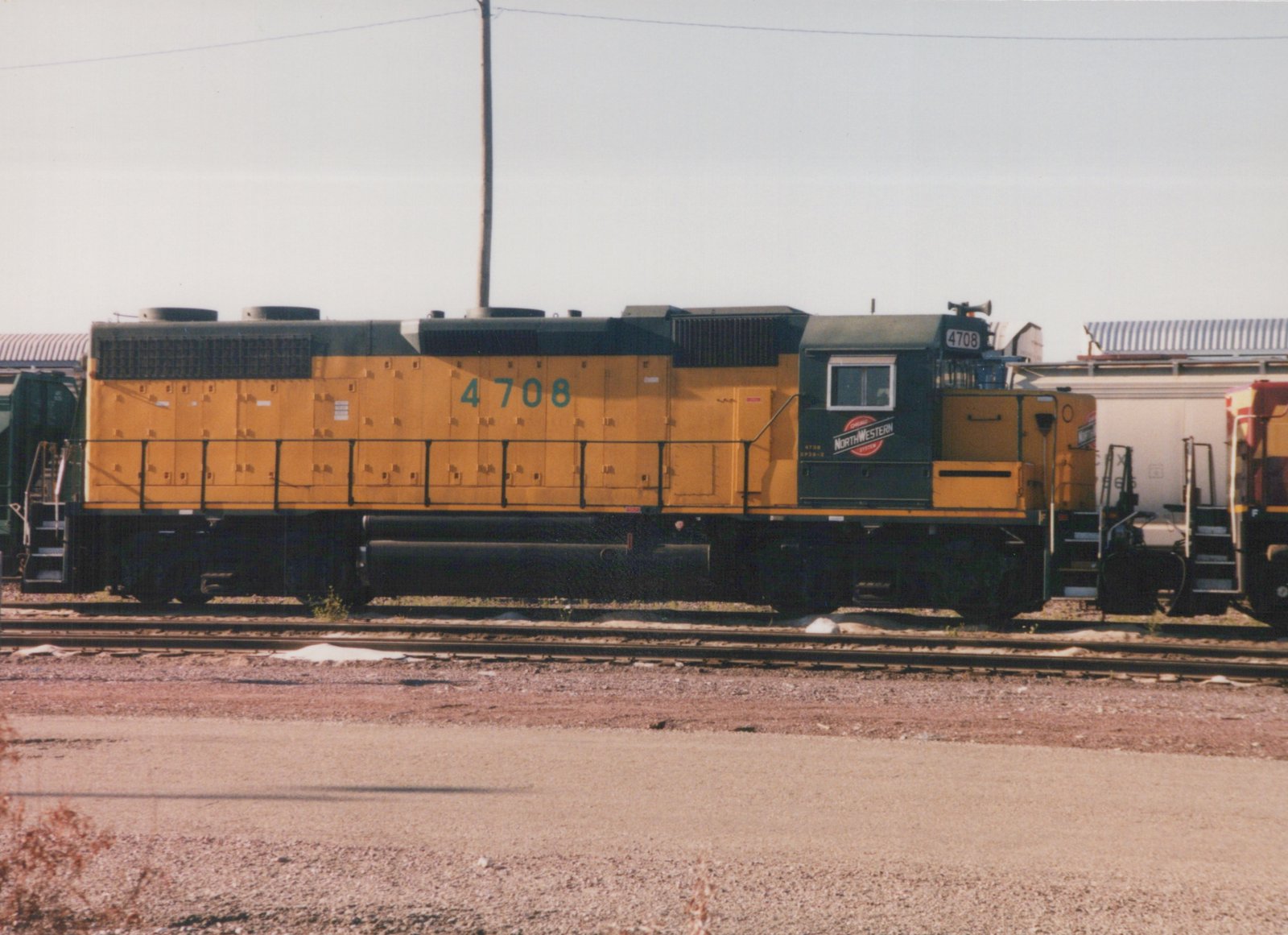 A side view of a yellow and black Chicago and North Western diesel locomotive, number 4708, standing on tracks in a gravel yard with a white boxcar and other freight cars nearby.
