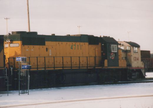 A side view of Chicago and North Western GP9 diesel locomotives 4711 and 1214 in a snow-covered yard under an overcast sky.