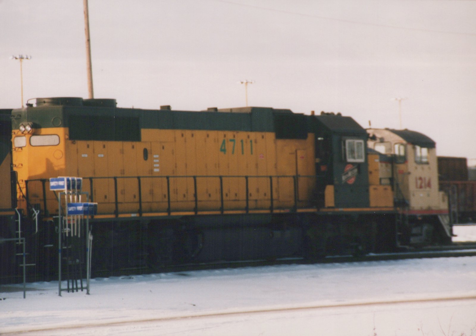 A side view of Chicago and North Western GP9 diesel locomotives 4711 and 1214 in a snow-covered yard under an overcast sky.