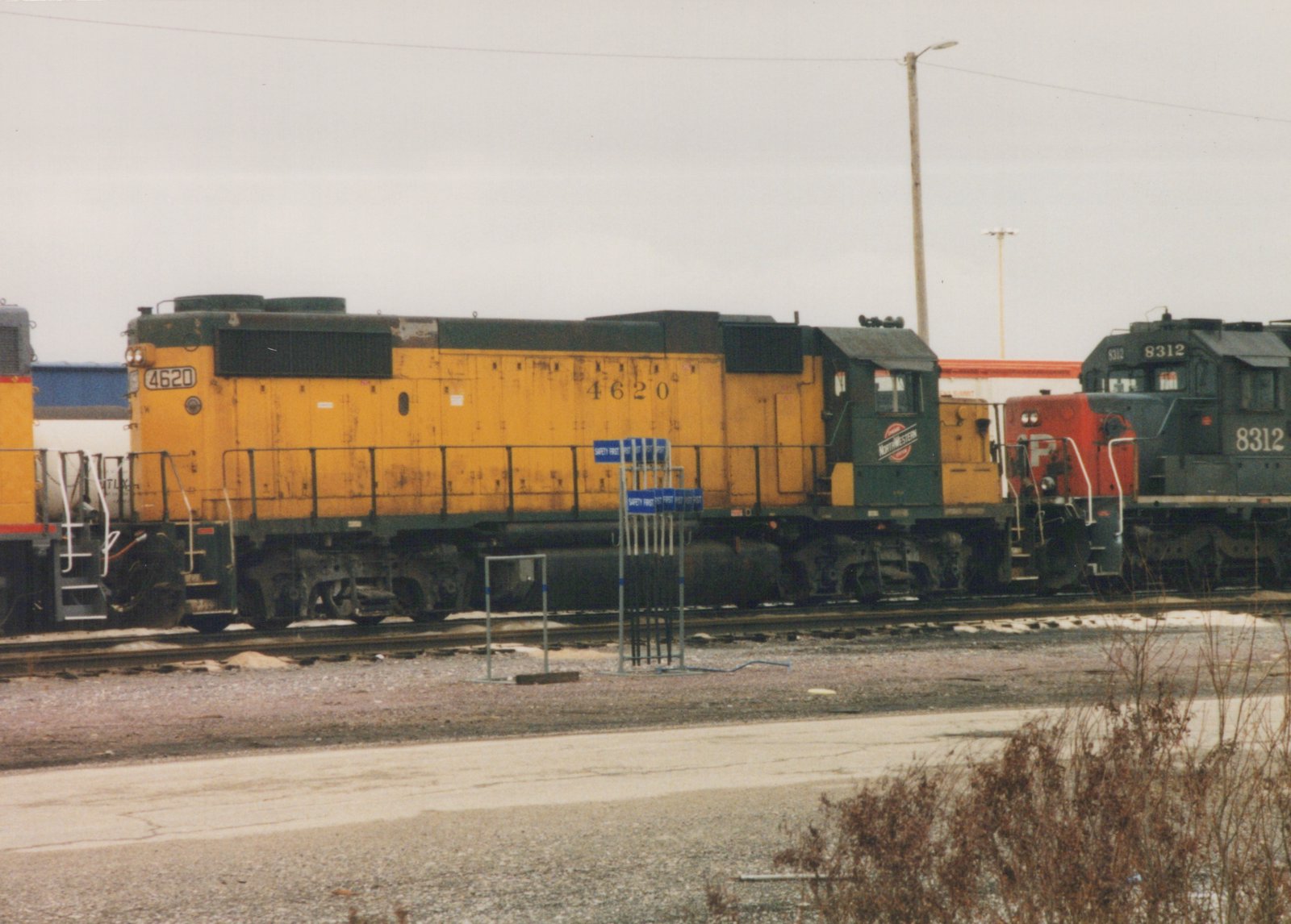 A yellow and green Chicago and North Western locomotive numbered 4620 is parked on tracks in a rail yard next to a dark locomotive with a red front and the number 8312, under a grey sky with patches of snow on the ground.
