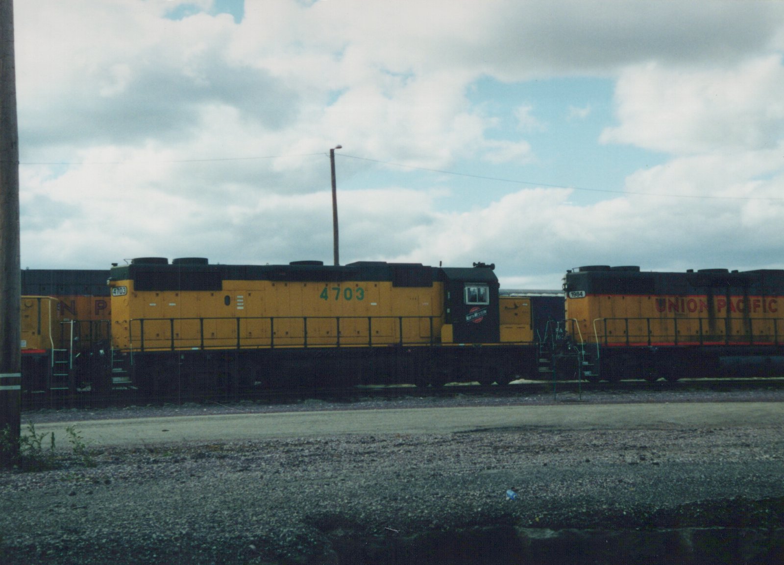Two yellow diesel locomotives, one Chicago and North Western #4703 and one Union Pacific #1984, are parked side by side in a gravel yard under an overcast sky.