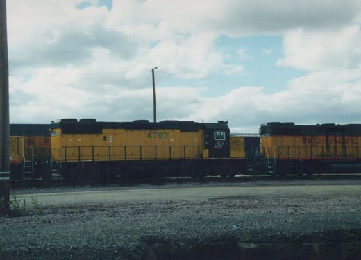 Two yellow diesel locomotives, one Chicago and North Western #4703 and one Union Pacific #1984, are parked side by side in a gravel yard under an overcast sky.