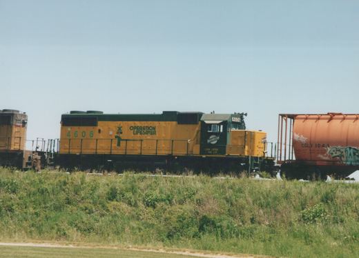 A yellow and green Chicago and North Western diesel locomotive, number 4606, with 'Operation Lifesaver' lettering on its side, is parked on a grassy railway line next to a red tank car.
