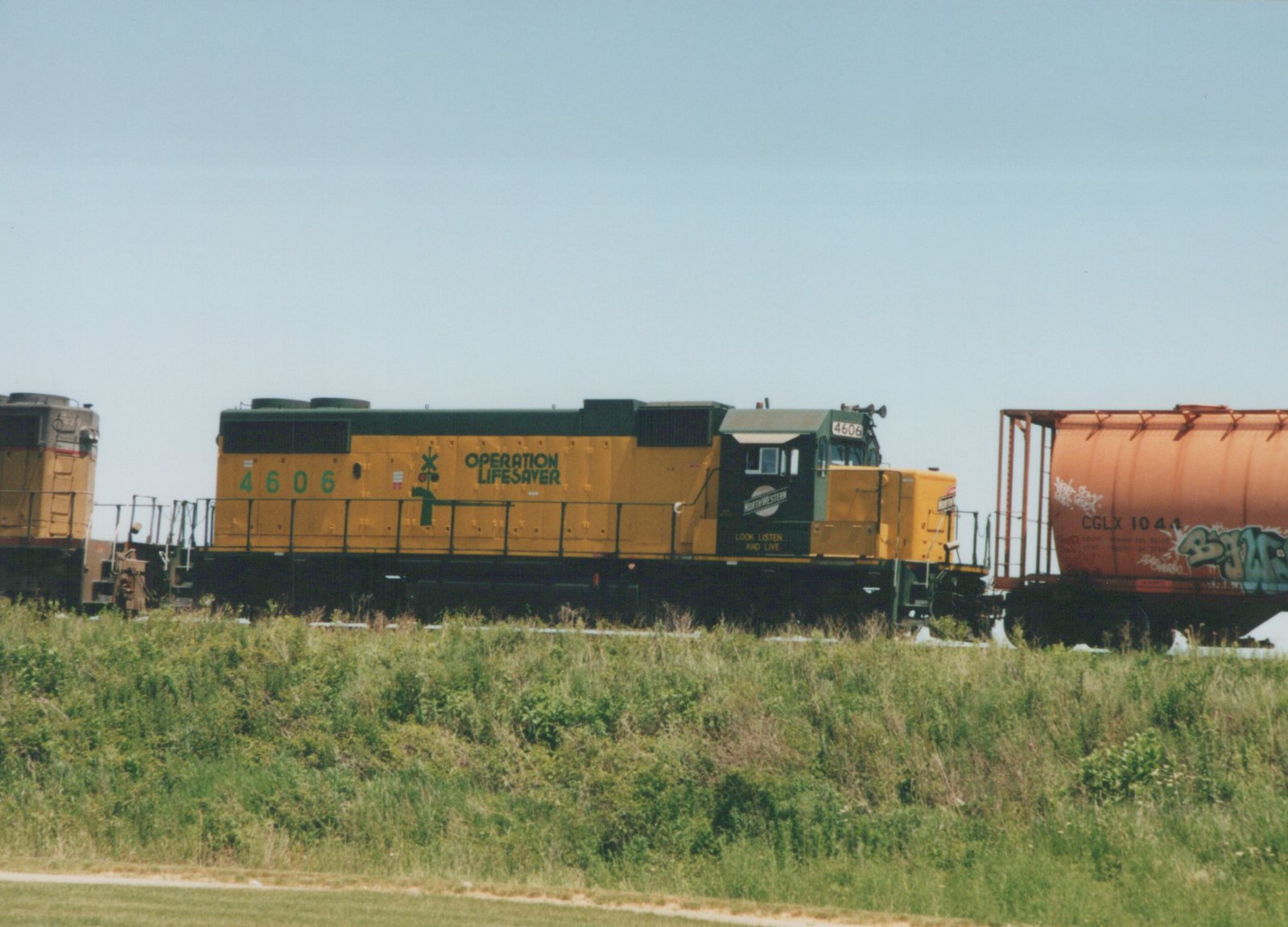 A yellow and green Chicago and North Western diesel locomotive, number 4606, with 'Operation Lifesaver' lettering on its side, is parked on a grassy railway line next to a red tank car.