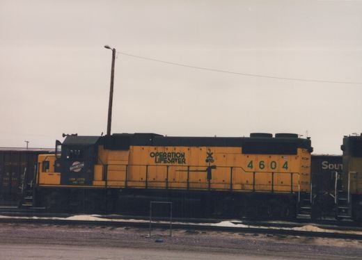 A yellow and black diesel locomotive with 'Operation Lifesaver' lettering sits on railroad tracks in a yard with snow on the ground, flanked by freight cars from Southern Pacific and Southern Railway.