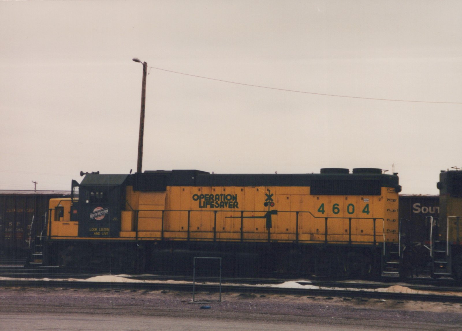 A yellow and black diesel locomotive with the number 4604 and "Operation Lifesaver" lettering is parked in a railroad yard next to dark freight cars under an overcast sky.