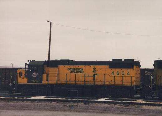 A yellow and black diesel locomotive with the number 4604 and "Operation Lifesaver" lettering is parked in a railroad yard next to dark freight cars under an overcast sky.