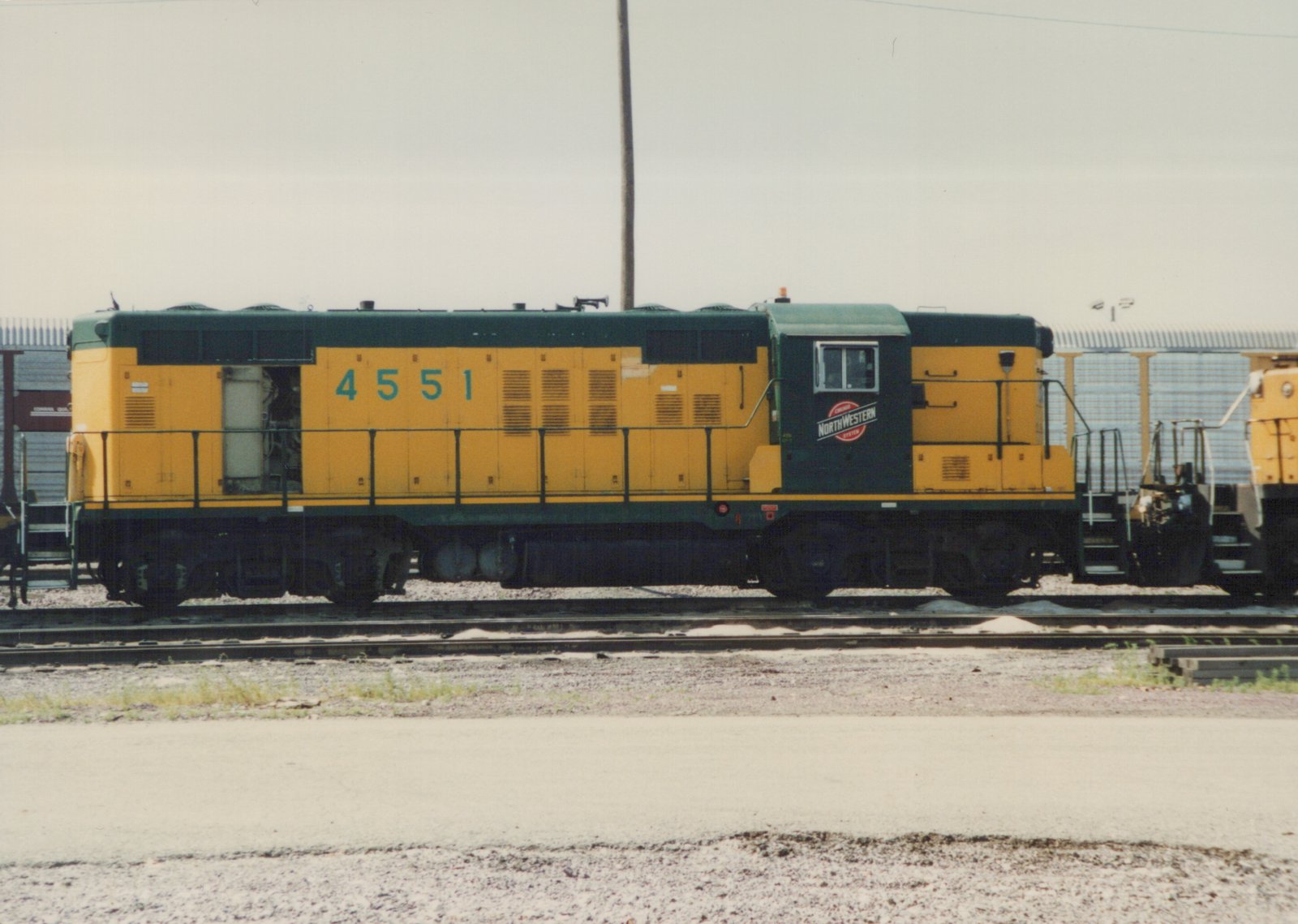 A side view of a yellow and green Chicago and North Western diesel locomotive, number 4551, sitting on tracks in a gravel yard under an overcast sky.