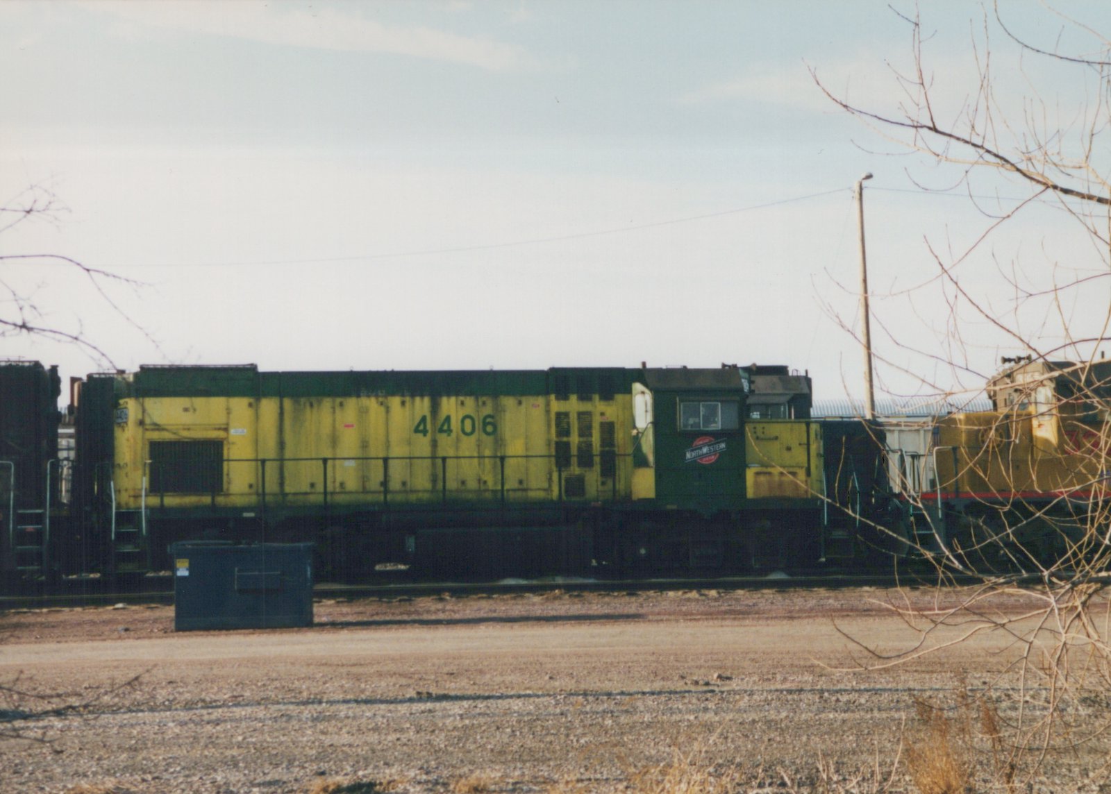 Side view of a yellow and green Chicago and North Western GP9 locomotive numbered 4406, parked in a gravel yard with bare trees and another locomotive partly visible behind.