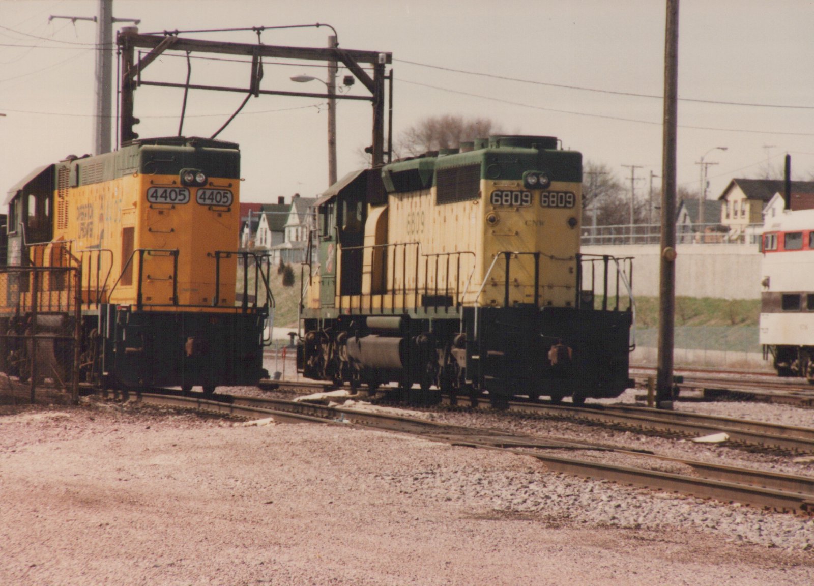 Two yellow and green diesel locomotives with numbers 4405 and 6809 sit on parallel tracks in a railroad yard under an overhead structure, with houses and a train in the background.