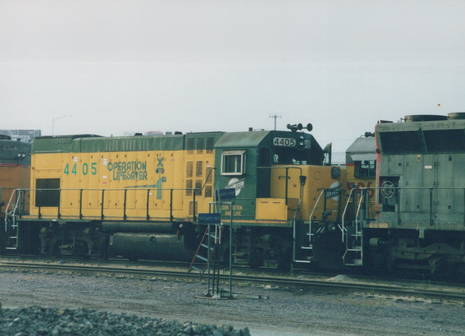 A yellow and green diesel locomotive with the number 4405 and 'Operation Lifesaver' text, parked on tracks in a yard with other locomotives nearby.