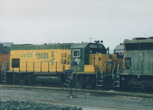 A yellow and green diesel locomotive with the number 4405 and 'Operation Lifesaver' text, parked on tracks in a yard with other locomotives nearby.