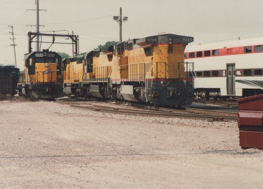A side view of two yellow diesel locomotives, one marked Chicago & North Western 4404 and the other Union Pacific 9368, standing in a gravel yard with a white, red, and brown Metra passenger car behind them under an overcast sky.
