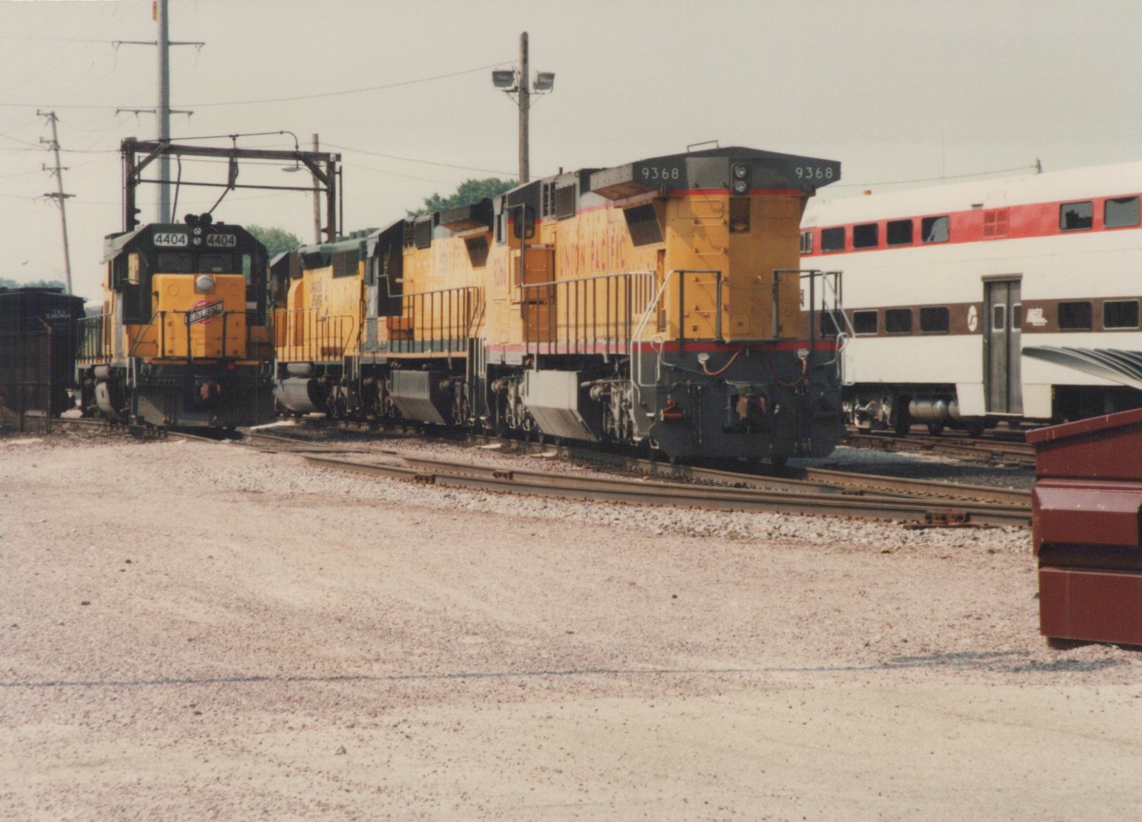 A side view of two yellow diesel locomotives, one marked Chicago & North Western 4404 and the other Union Pacific 9368, standing in a gravel yard with a white, red, and brown Metra passenger car behind them under an overcast sky.
