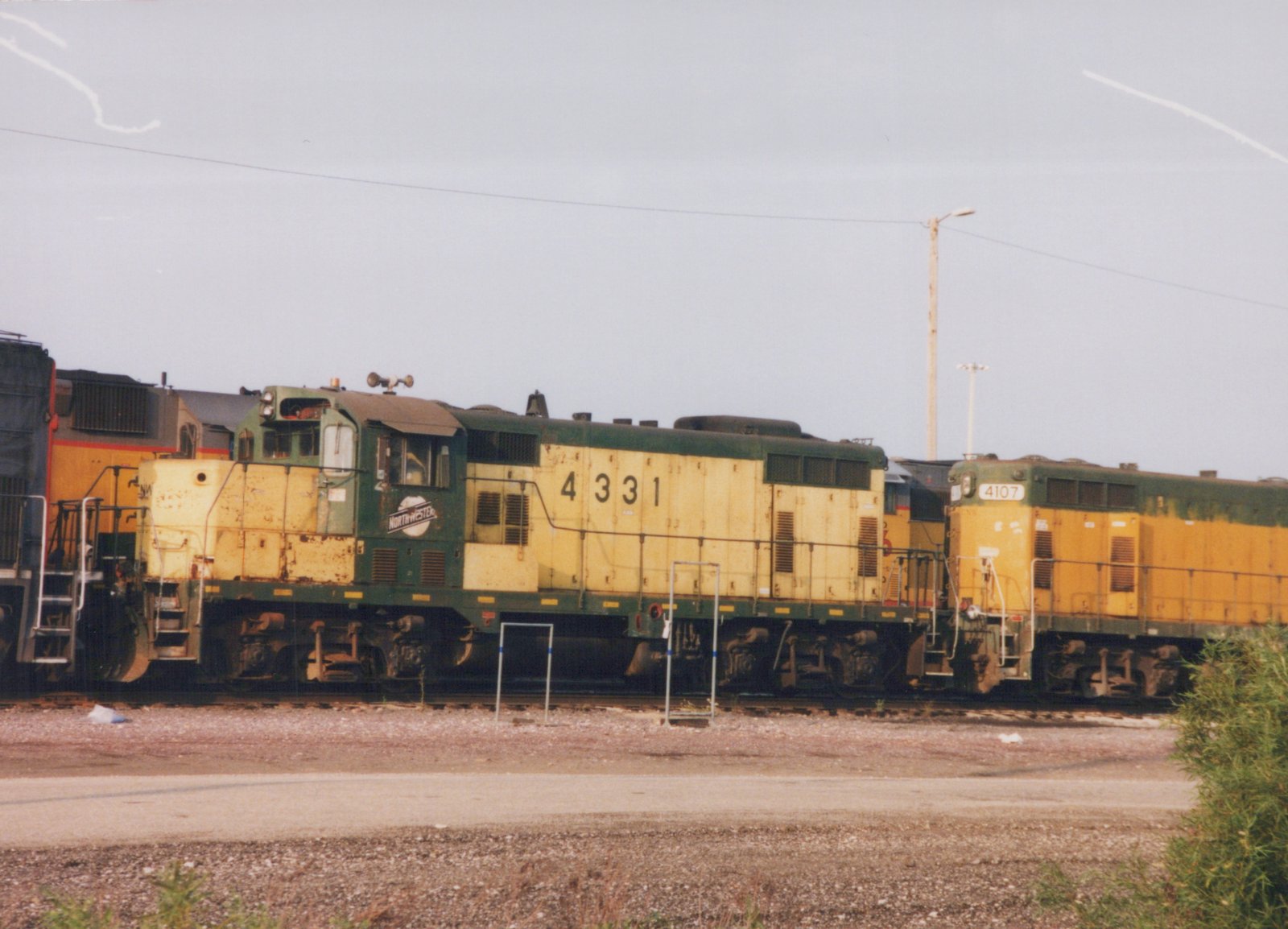 Two yellow and green diesel locomotives, one marked 4331 and the other 4107, sit on tracks in a gravel yard under an overcast sky.