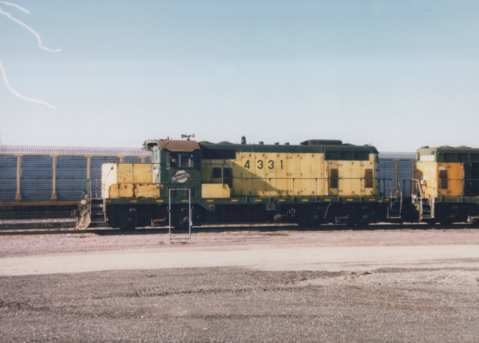 A side view of two green and yellow Chicago and North Western diesel locomotives, numbered 4331 and 4107, parked on tracks in a gravel yard under a clear blue sky.