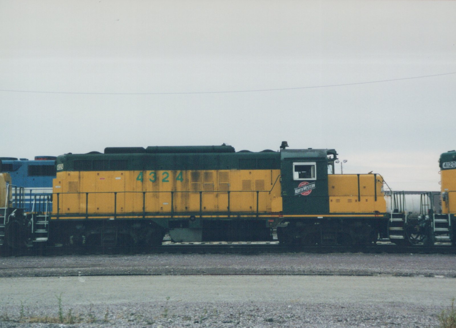 Side view of a yellow and green Chicago and North Western GP9 diesel locomotive, number 4324, parked in a gravel yard with another locomotive behind it.
