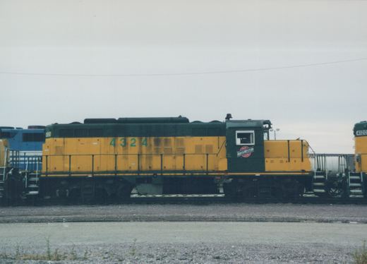 Side view of a yellow and green Chicago and North Western GP9 diesel locomotive, number 4324, parked in a gravel yard with another locomotive behind it.