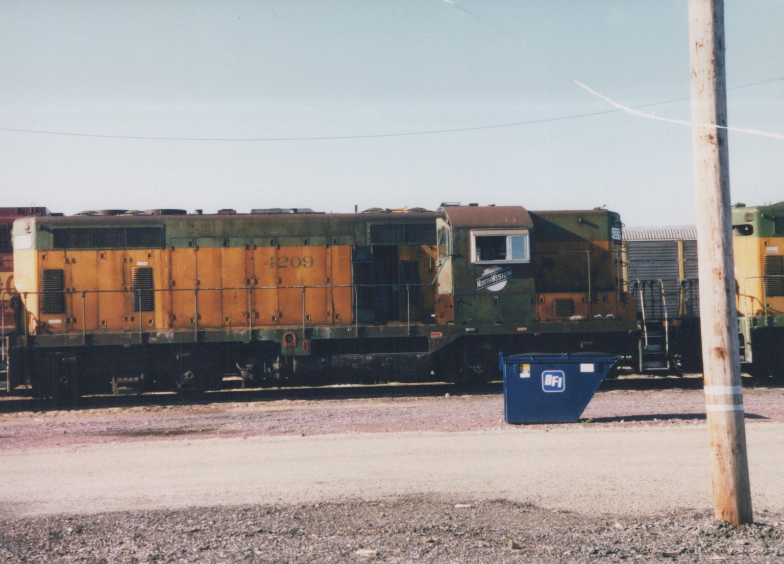 Side view of a Chicago and North Western GP9 diesel locomotive, number 4209, in orange and green, parked on a track in a yard under a clear sky.