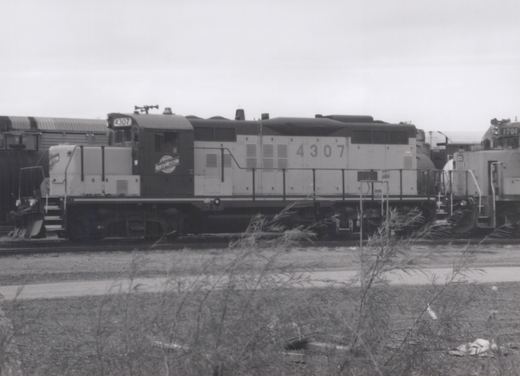 A side view of a black and gray diesel locomotive with the number 4307 and 'NORTH WESTERN' on its cab, parked in a rail yard with overgrown weeds in the foreground.