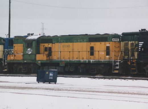 A green and yellow Chicago and North Western GP9 locomotive numbered 4195 is parked on snowy tracks in a rail yard, with a blue BFI dumpster in the foreground.