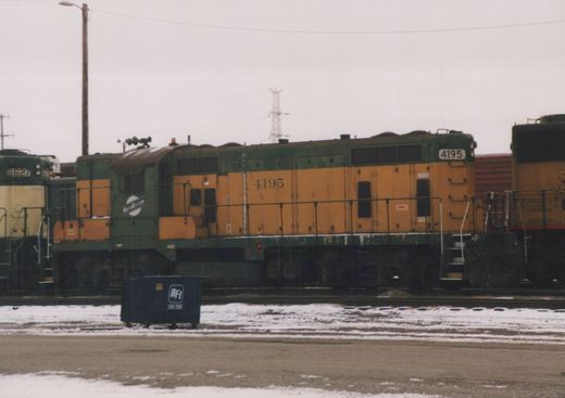 A green and orange Chicago and North Western diesel locomotive, number 4195, sits on tracks in a snowy yard next to another locomotive and a blue dumpster.