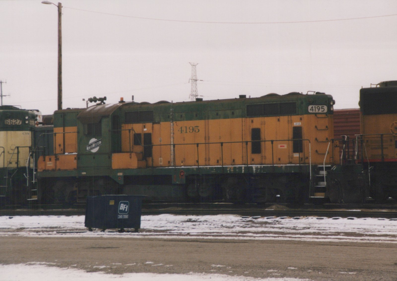 A green and orange Chicago and North Western diesel locomotive, number 4195, sits on tracks in a snowy yard next to another locomotive and a blue dumpster.