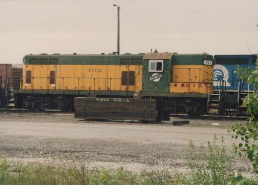 A side view of a green and yellow Chicago and North Western GP9 diesel locomotive, number 4161, parked in a gravel lot with a blue Burlington Northern locomotive behind it.
