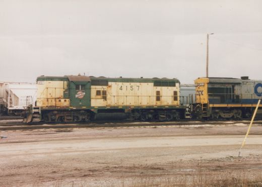 Side view of a green and cream Chicago and North Western locomotive, number 4157, coupled to a blue and yellow CSX locomotive in a muddy rail yard under a gray sky.