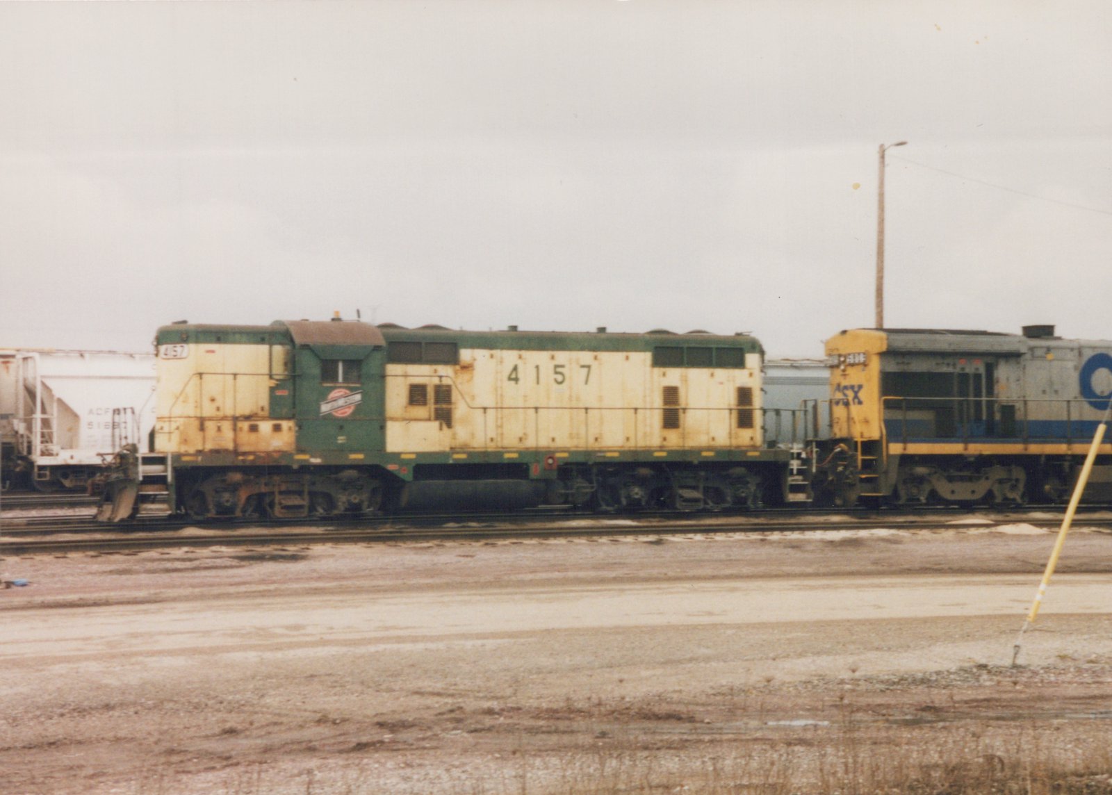 Side view of a green and cream Chicago and North Western locomotive, number 4157, coupled to a blue and yellow CSX locomotive in a muddy rail yard under a gray sky.