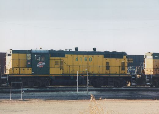 A side view of a yellow and green Chicago and North Western diesel locomotive, number 4160, in a rail yard with other equipment in the background.