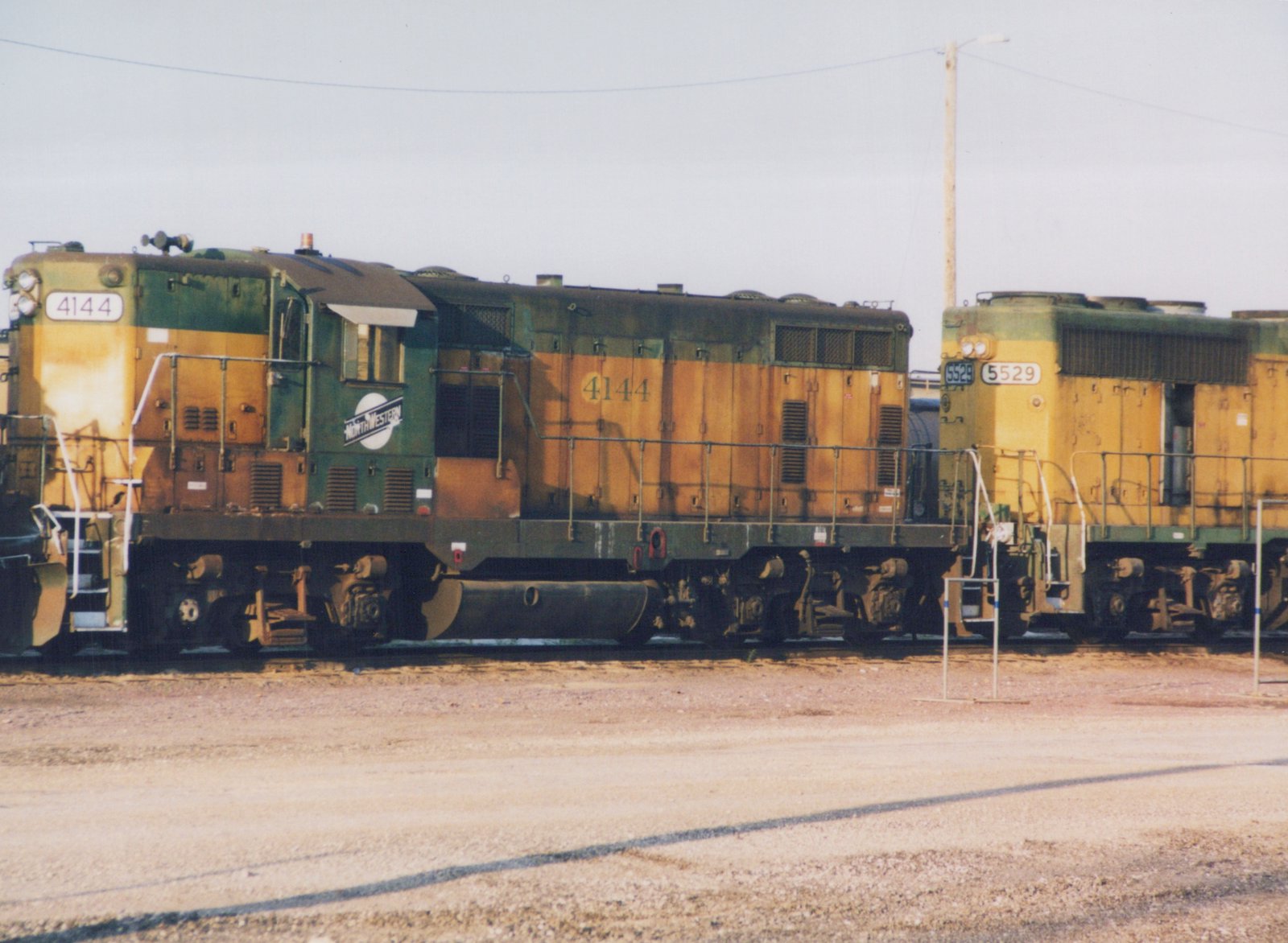 Two green and yellow Chicago and North Western diesel locomotives, one numbered 4144 and the other 5529, are parked next to each other in a gravel yard under an overcast sky.