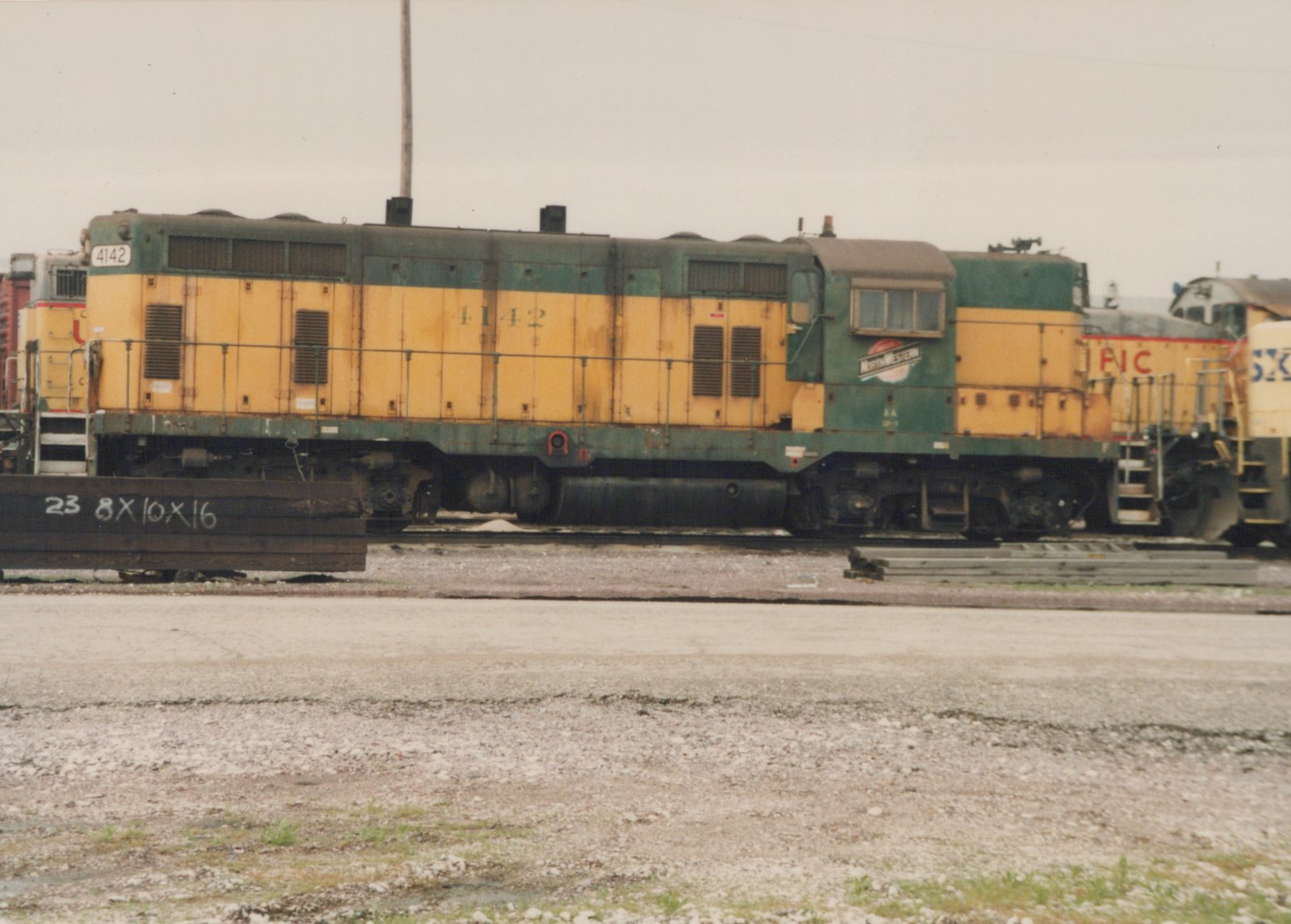 A side view of a yellow and green diesel locomotive with the number 4142 and a North Western herald on its cab, standing on tracks in a gravel yard under an overcast sky.