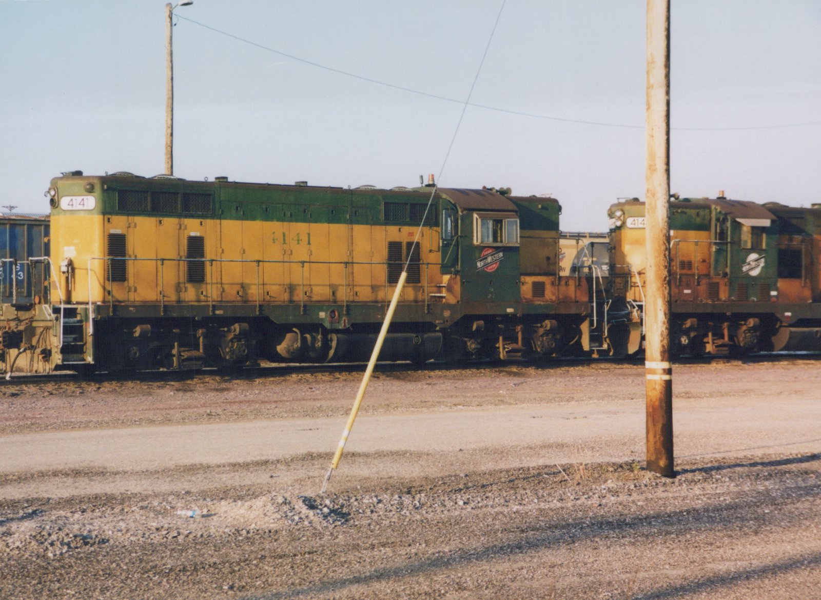 Two Chicago and North Western diesel locomotives, 4141 and 414?, parked in a gravel yard under a clear sky.