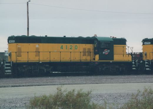 Side view of a yellow and green Chicago and North Western diesel locomotive numbered 4120, with 4190 partially visible behind it, both in a gravel yard under an overcast sky.