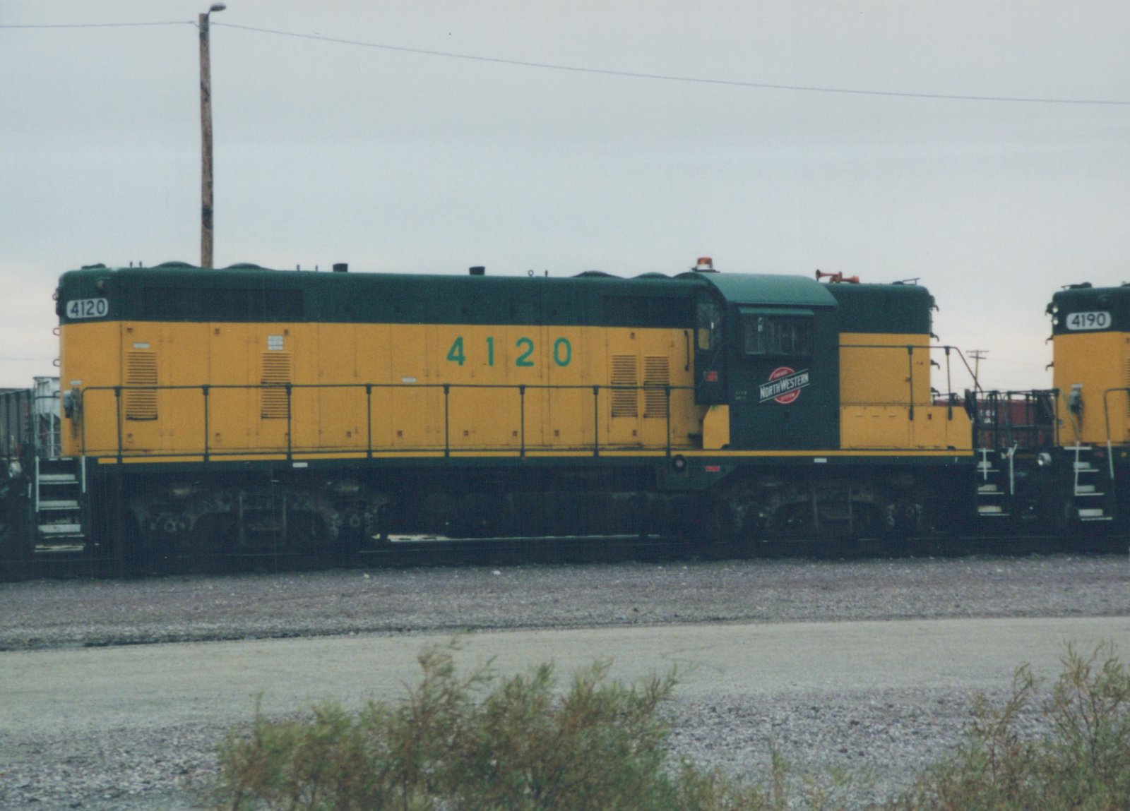 Side view of a yellow and green Chicago and North Western diesel locomotive numbered 4120, with 4190 partially visible behind it, both in a gravel yard under an overcast sky.