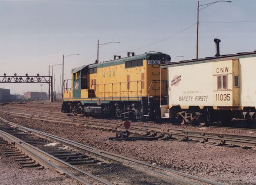 A yellow and green Chicago and North Western GP7 locomotive numbered 4108 is coupled to a matching caboose numbered 11035, parked on parallel railroad tracks in a yard with signal bridges in the background.
