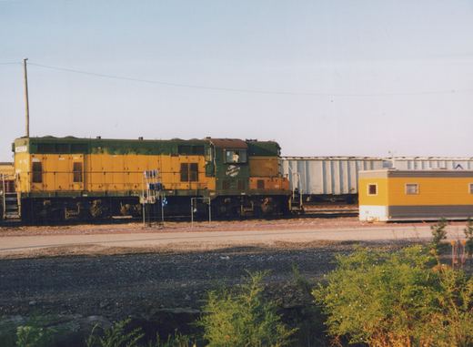 A green and yellow Chicago and North Western diesel locomotive, number 4107, sits on tracks in a rail yard with freight cars and a yellow trailer nearby under a clear sky.