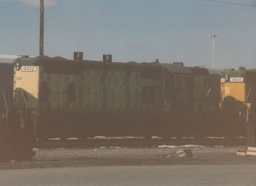 Two green and yellow Chicago and North Western diesel locomotives, numbered 4102 and 4114, sit side by side in a gravel yard under a clear sky, with patches of snow visible on the ground.