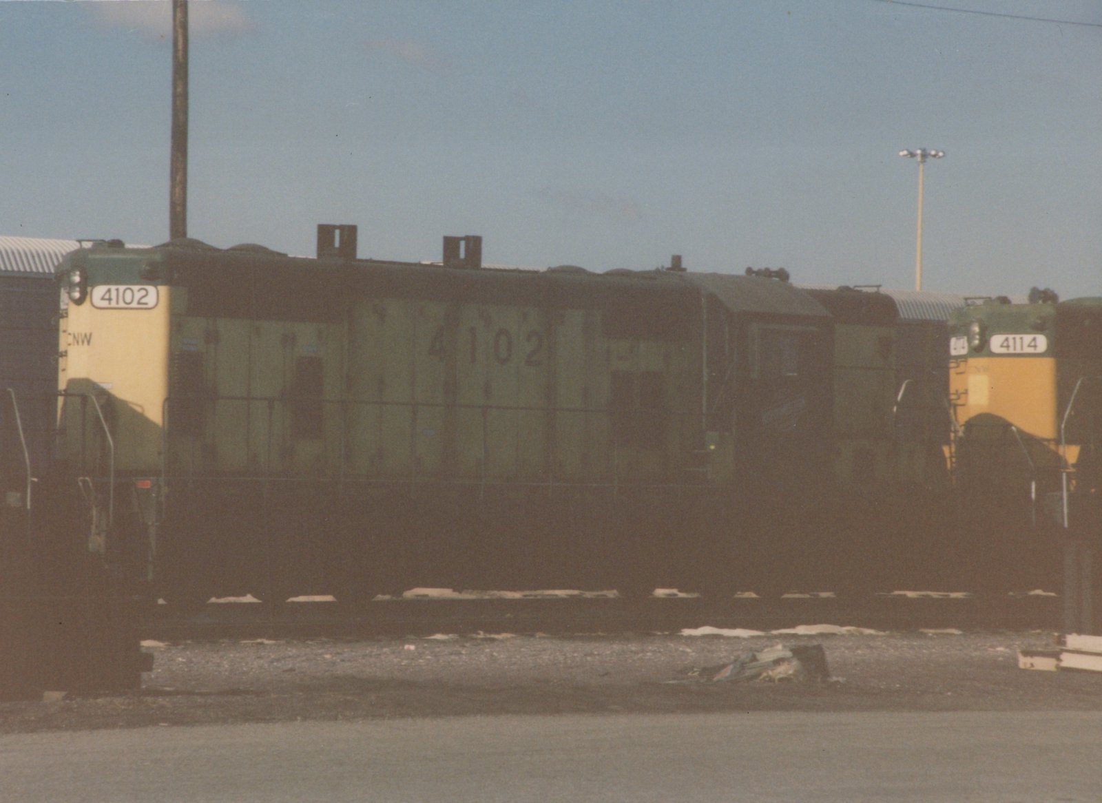 Two green and yellow Chicago and North Western diesel locomotives, numbered 4102 and 4114, sit side by side in a gravel yard under a clear sky, with patches of snow visible on the ground.