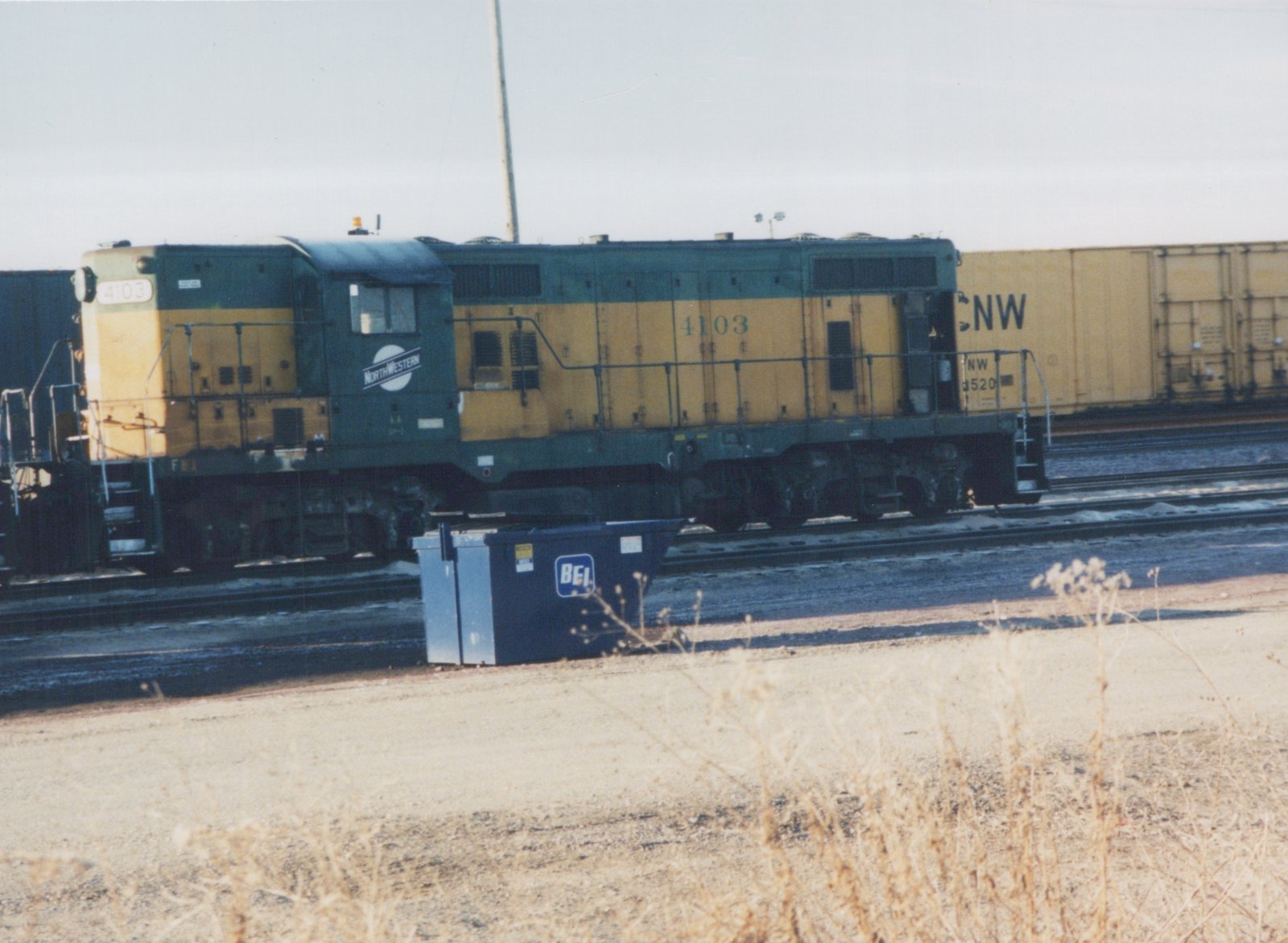 A green and yellow Chicago and North Western diesel locomotive, number 4103, stands on the tracks in a yard with a freight car behind it and a blue dumpster in the foreground.